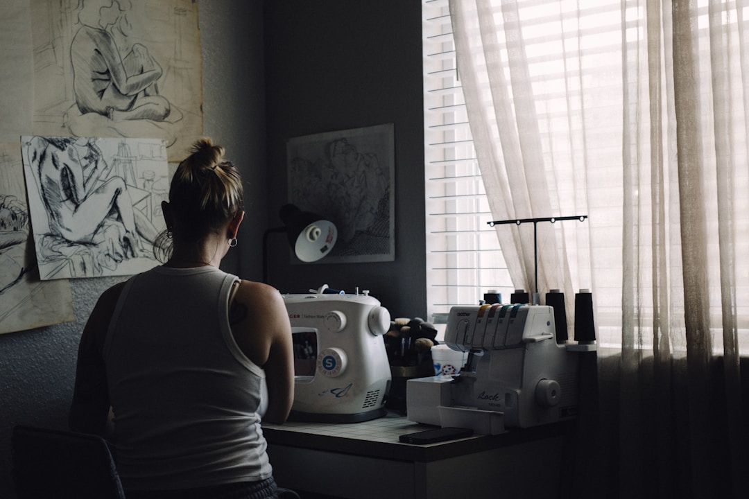 Black woman at a sewing machine, back to camera, in a cozy creative studio with sketches and fabric on the walls